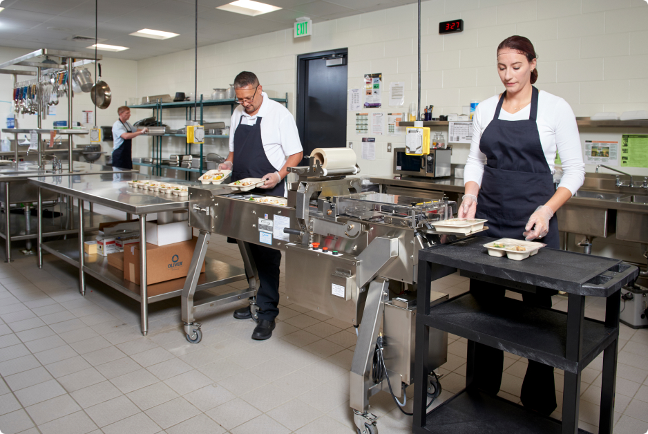 Workers operating food tray sealing equipment in a commercial kitchen.